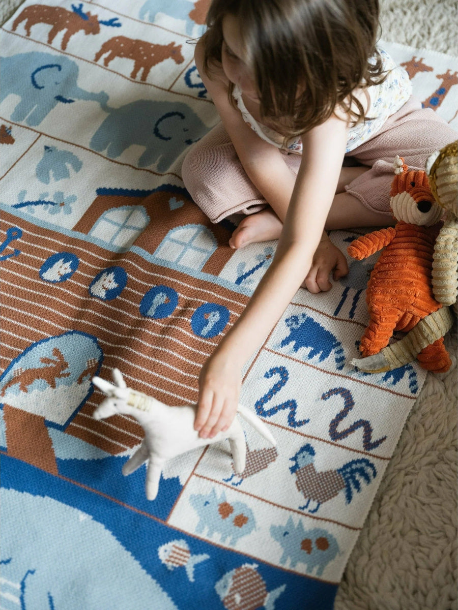 Young toddler playing on a soft Biquette Ark Midi knit blanket designed by Amelie Mancini featuring colorful animal illustrations.
