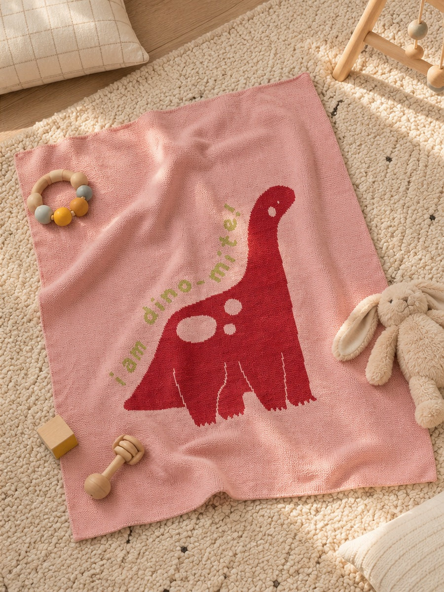 Pink Biquette Mini knit blanket designed by Ellie Hyde featuring a red dinosaur design, laid on the floor with soft folds and baby toys for infant playtime.