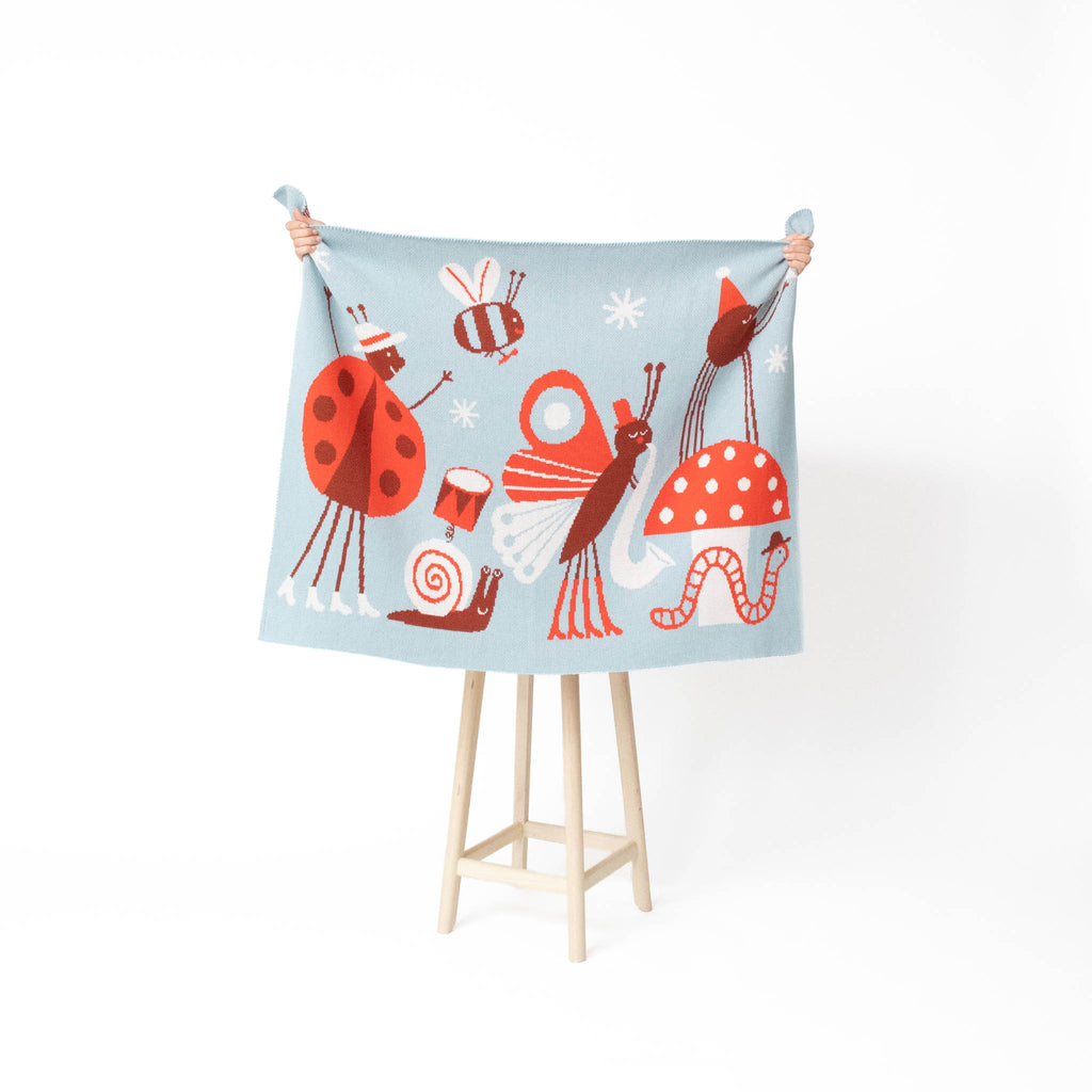 A light blue fabric blanket or throw featuring whimsical cartoon bugs and mushrooms, held up by a person's hands on a wooden stand against a white background.