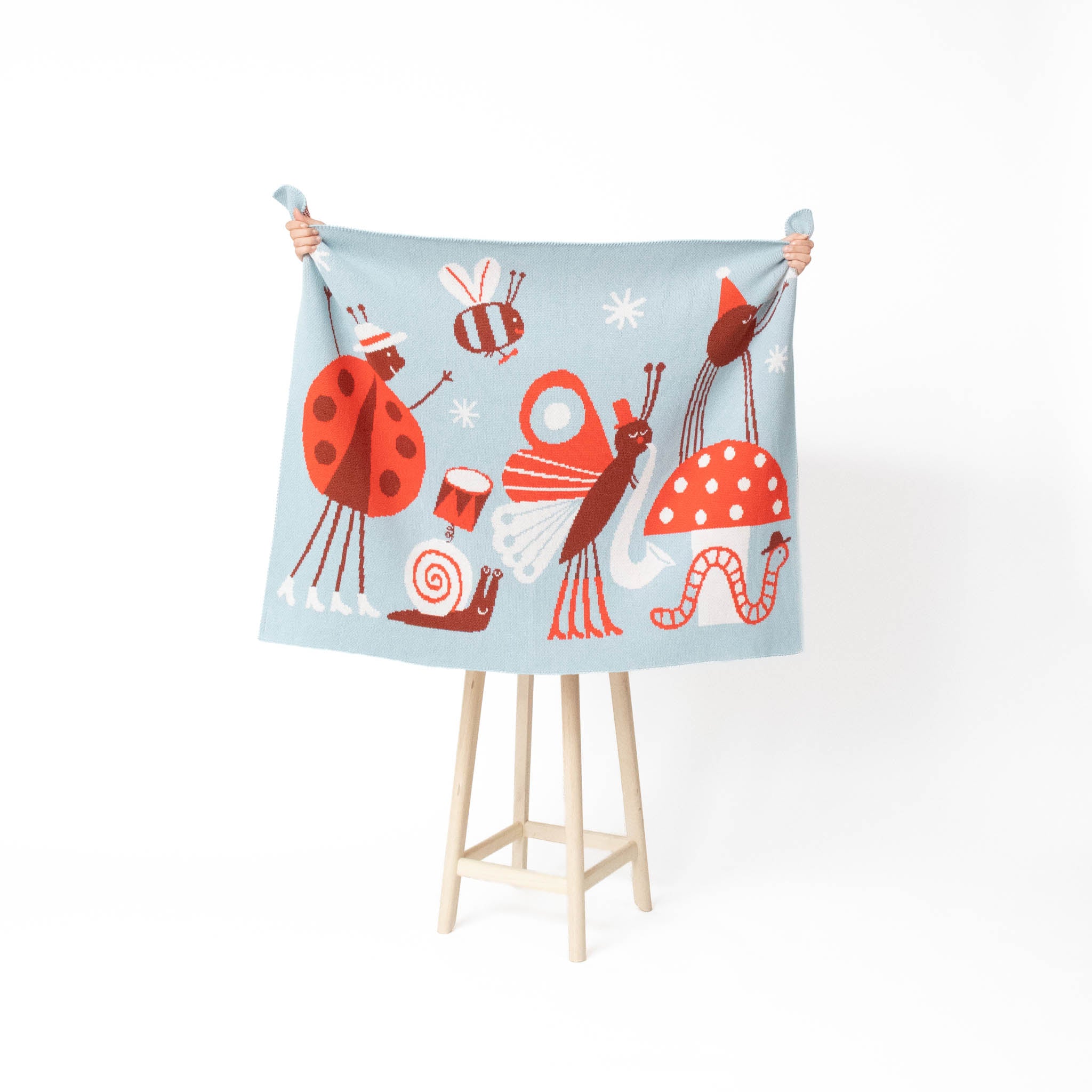 A light blue fabric blanket or throw featuring whimsical cartoon bugs and mushrooms, held up by a person's hands on a wooden stand against a white background.