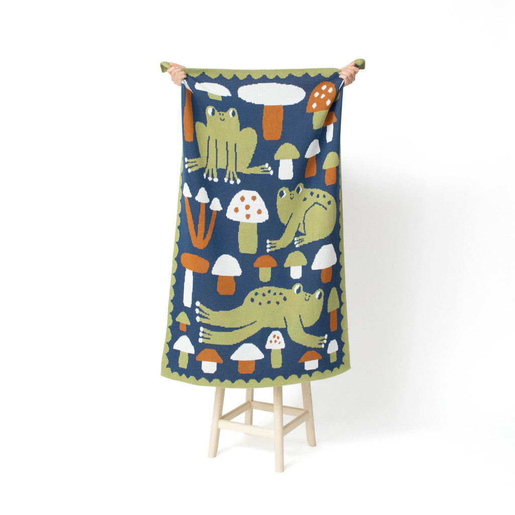 A rectangular knit blanket, held up by two hands on a small wooden stand against a plain white background. The textile features a whimsical pattern of green frogs and orange and white mushrooms on a dark navy blue base, with a green scalloped border.