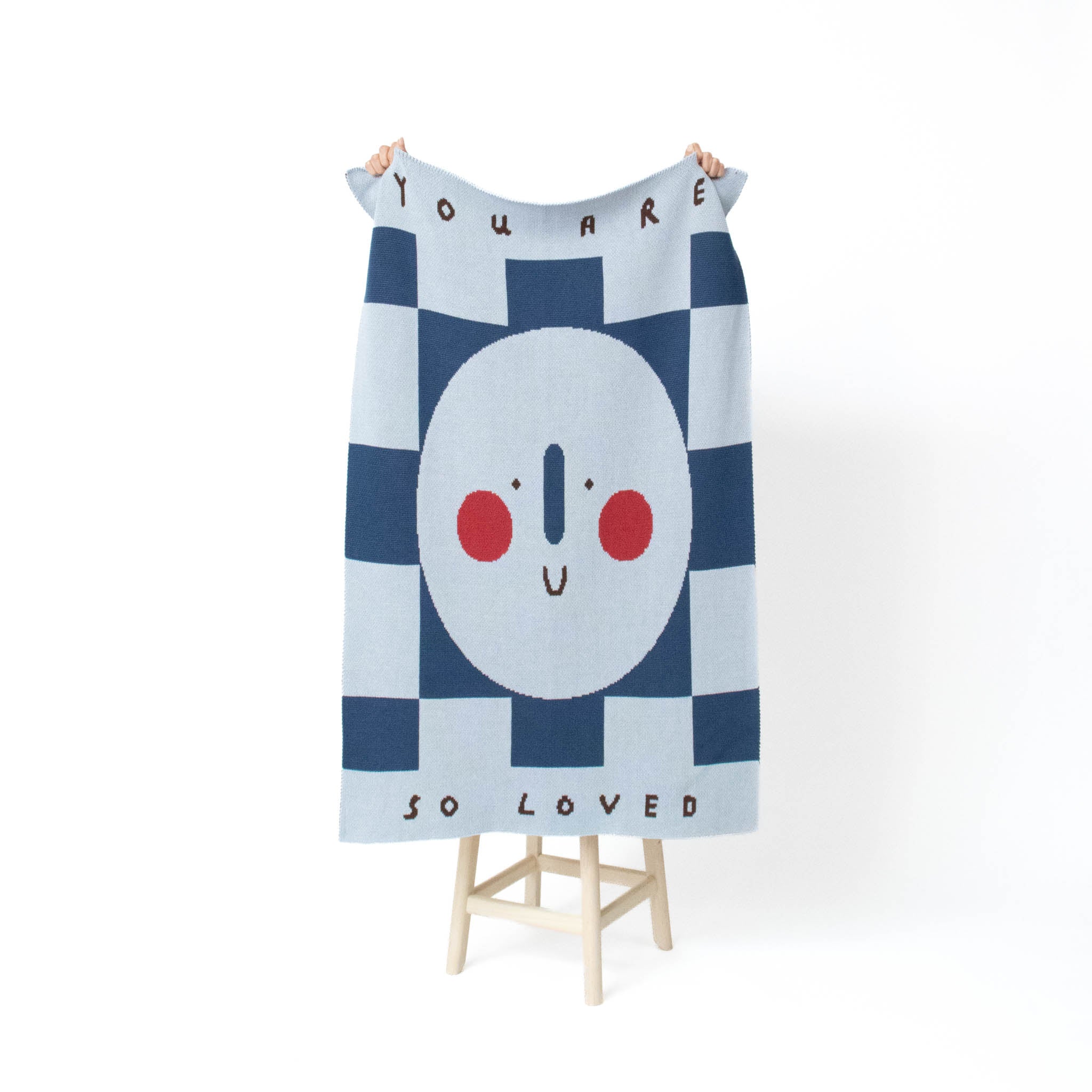 A knitted blanket with a navy and light blue checkerboard pattern, featuring a central white circle with a minimalist face and red cheeks, held up by hands on a small wooden stand against a white background.